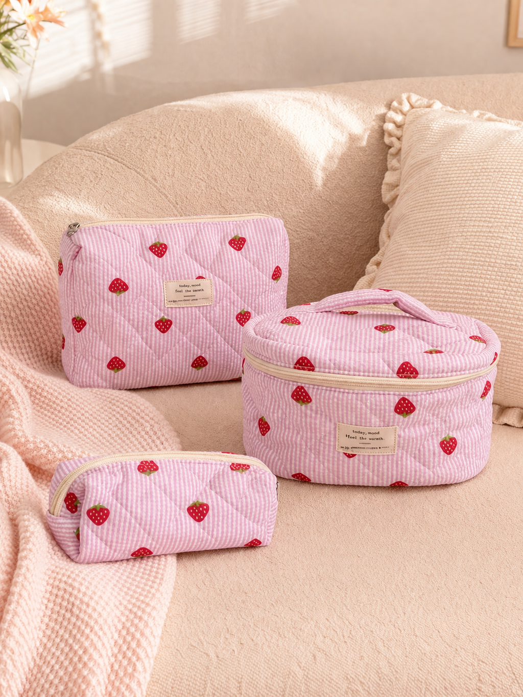 Set of pink cosmetic bags with strawberry pattern on a beige couch.